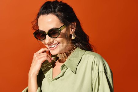 Téléchargez les photos : Une femme élégante dans une chemise verte et des lunettes de soleil frappant une pose dans un cadre de studio sur un fond orange. - en image libre de droit