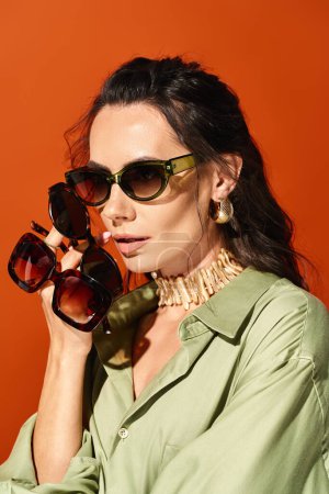 Foto de Una mujer elegante con una camisa verde y gafas de sol posa en un estudio sobre un fondo naranja, encarnando la moda veraniega. - Imagen libre de derechos