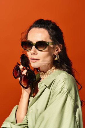 A stylish woman exudes summertime vibes wearing a green shirt and fashionable sunglasses against an orange background.