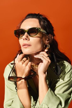 A stylish woman with sunglasses poses in a green shirt in a studio against an orange background, exuding summertime fashion vibes.