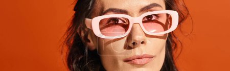 Téléchargez les photos : Une femme à la mode avec une chemise blanche et des lunettes de soleil roses pose sur un fond de studio orange, respirant le charme de l'été. - en image libre de droit