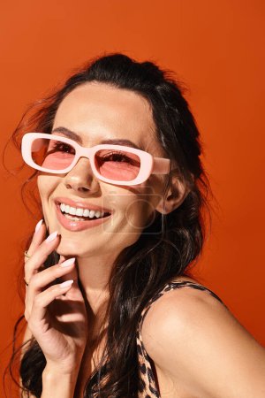 A stylish woman is striking a pose while wearing pink sunglasses against an orange studio backdrop.