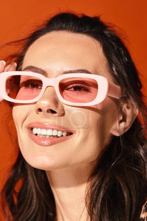 A pretty woman in pink sunglasses smiles brightly in a studio, showcasing summertime fashion on an orange background.