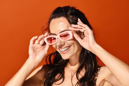 Una mujer elegante con una sonrisa brillante que se pone gafas de sol rosadas en un entorno de estudio con un fondo naranja, exudando vibraciones de moda de verano.