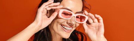 A lovely woman beams while sporting pink heart-shaped sunglasses against an orange studio backdrop.