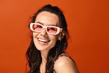 Foto de Una mujer con estilo irradia alegría mientras se pone gafas de sol rosadas con un vibrante telón de fondo naranja, exudando vibraciones de moda de verano. - Imagen libre de derechos