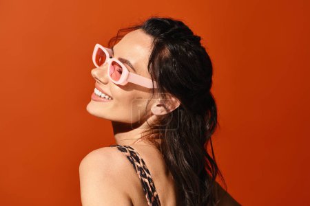 Téléchargez les photos : Une femme à la mode respire la confiance dans les lunettes de soleil roses et une robe imprimée léopard sur fond orange vif. - en image libre de droit