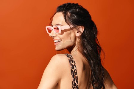 Téléchargez les photos : Une femme à la mode dégage des vibrations estivales dans un haut imprimé léopard, accessoirisé avec des lunettes de soleil roses, sur un fond orange. - en image libre de droit