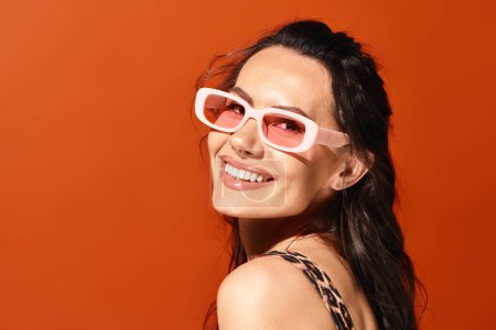 Foto de Una mujer con estilo en gafas de sol de color rosa posa con confianza delante de una pared naranja vibrante, encarnando la moda de verano. - Imagen libre de derechos