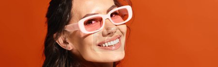 A stylish woman donning pink sunglasses smiles brightly at the camera in a vibrant studio setting against an orange background.