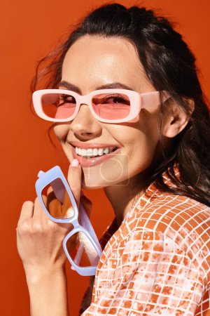 Foto de Una mujer elegante con gafas de sol rosas emana vibraciones de verano mientras sonríe con un fondo naranja. - Imagen libre de derechos