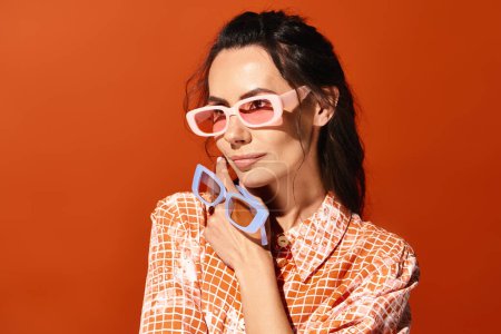 A fashionable woman poses in pink sunglasses and a colorful patterned shirt on a vibrant orange background.