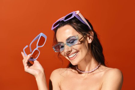A stylish woman in sunglasses holding up a pair of glasses in a studio against an orange background, showcasing summertime fashion.
