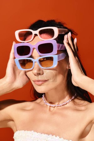 Foto de Una mujer impresionante con un vestido blanco y gafas de sol púrpura posa en un estudio sobre un fondo naranja, encarnando la moda veraniega. - Imagen libre de derechos