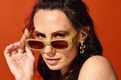 A fashionable woman posing in a studio, wearing bright yellow sunglasses against a vibrant orange backdrop, exuding summertime vibes. magic mug #711225446