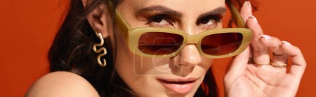 Téléchargez les photos : Une femme à la mode dégage des vibrations estivales dans un studio, berçant des lunettes de soleil jaune vif sur fond orange. - en image libre de droit