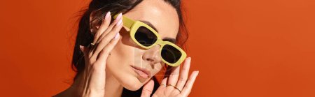 Eine stilvolle Frau mit gelber Sonnenbrille posiert mit zart ins Gesicht gelegten Händen und strahlt auf orangefarbenem Studiohintergrund Zuversicht und Sommermode aus..