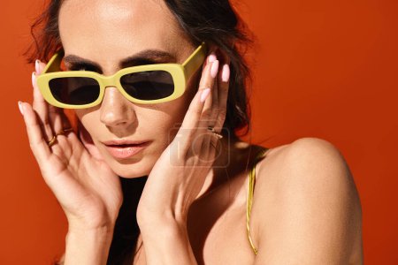 A fashionable woman wearing yellow sunglasses, hands on face, in studio on orange background.