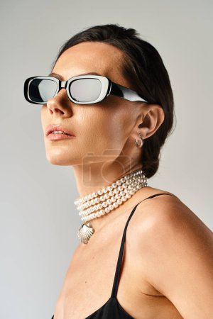 Eine stilvolle Frau ziert eine Sonnenbrille und Perlen und strahlt Eleganz und Raffinesse vor grauem Hintergrund aus.