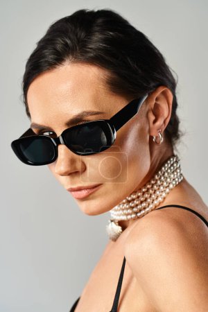 Une femme à la mode avec des perles autour du cou frappant une pose dans des lunettes de soleil sur un fond gris.
