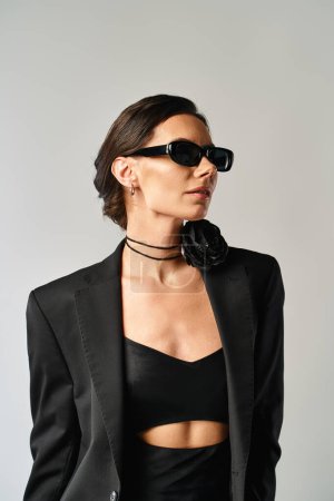 Eine modische Frau strahlt in schwarzem Anzug und Sonnenbrille Zuversicht aus und posiert in einem Studio vor grauem Hintergrund.
