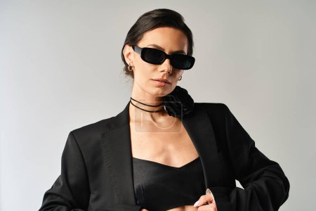 Foto de Una mujer con estilo en un traje negro y gafas de sol emana confianza y misterio en un estudio con un fondo gris. - Imagen libre de derechos