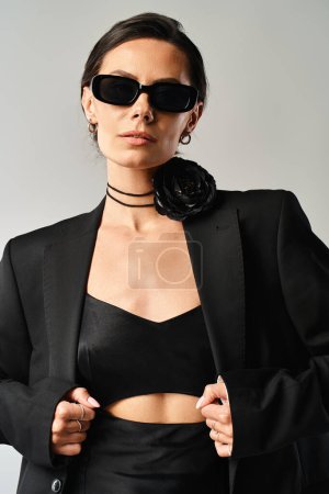 A stylish woman exudes confidence in a black suit and sunglasses in a professional photoshoot on a grey background.