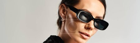 Téléchargez les photos : Une femme à la mode respire la confiance dans une robe noire et des lunettes de soleil, posant sur un fond gris dans un studio. - en image libre de droit
