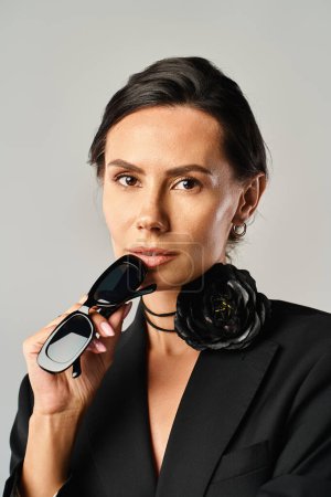Foto de Una mujer con estilo en un traje negro sostiene unas gafas de sol, mostrando elegancia y sofisticación en un estudio sobre un fondo gris. - Imagen libre de derechos