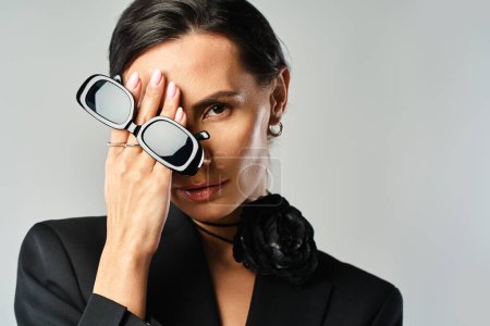 Une femme élégante en costume noir tient en toute confiance une paire de lunettes dans un cadre studio avec un fond gris.