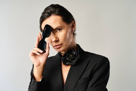A stylish woman in a suit confidently holds a pair of black sunglasses in a studio setting