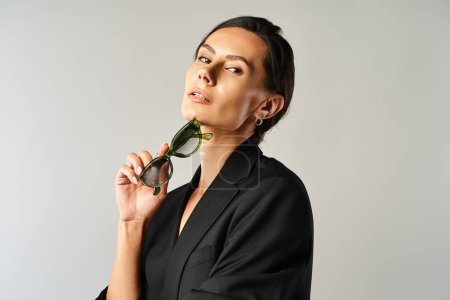 Eine stilvolle Frau setzt sich in einem Studio vor grauem Hintergrund eine Brille auf das Gesicht.