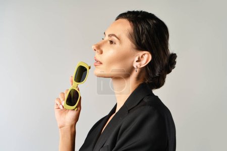 Una mujer de moda sostiene alegremente un par de gafas de sol de color amarillo brillante contra un fondo gris.