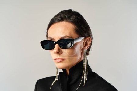 A fashionable woman with sunglasses poses in a black jacket against a grey studio backdrop.