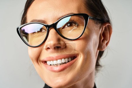 A fashionable woman, wearing glasses, smiles brightly at the camera in a studio setting against a grey background.