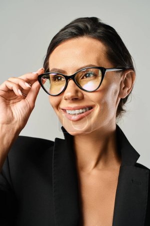 A fashionable woman with sleek sunglasses and a trendy black jacket poses in a studio against a grey background.