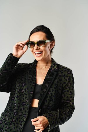 A fashionable woman in a black jacket and sunglasses poses in a studio setting against a grey background.