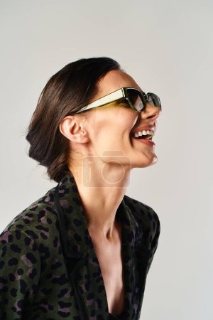 A stylish woman confidently showcases a leopard print shirt and trendy sunglasses in a studio setting against a grey background.