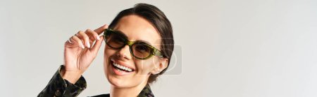 Eine stilvolle Frau strahlt Zuversicht aus, rockt schicke Sonnenbrillen und strahlt in einem Studio vor grauem Hintergrund ein strahlendes Lächeln aus..