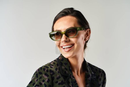 Una mujer elegante con gafas de sol emana felicidad en un estudio sobre un fondo gris.