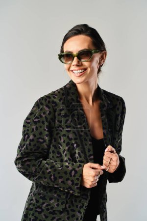 A fashionable woman poses in a stylish jacket and sunglasses against a grey studio backdrop.