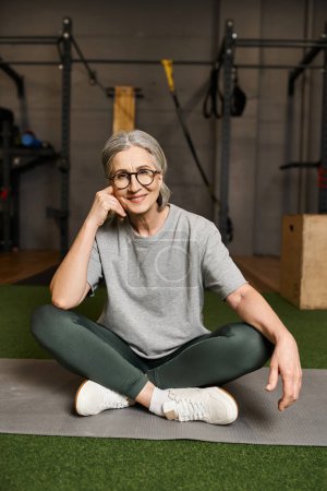 freudige reife Frau in bequemer Sportbekleidung sitzt mit überkreuzten Beinen im Fitnessstudio und lächelt in die Kamera
