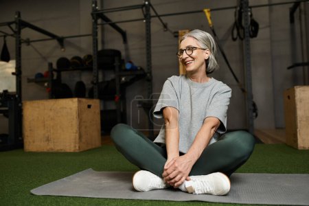 Photo for Appealing jolly mature woman in sportswear sitting on floor with crossed legs and looking away - Royalty Free Image