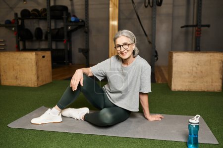 Téléchargez les photos : Positive belle femme mature en vêtements de sport avec des lunettes assises sur le sol et souriant à la caméra - en image libre de droit