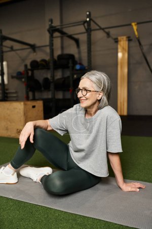Foto de Atractiva mujer madura alegre en camiseta gris con gafas sentadas en el suelo y mirando hacia otro lado - Imagen libre de derechos
