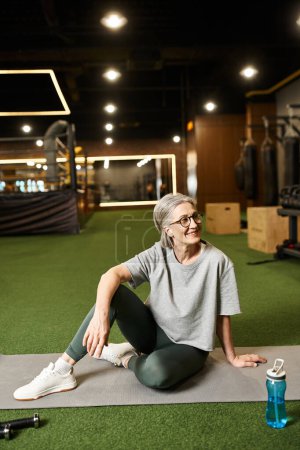 Photo for Attractive happy mature woman with gray hair and glasses sitting on floor in gym and looking away - Royalty Free Image