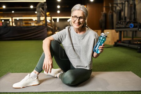 Photo for Good looking jolly mature sportswoman with glasses holding water bottle and smiling at camera - Royalty Free Image
