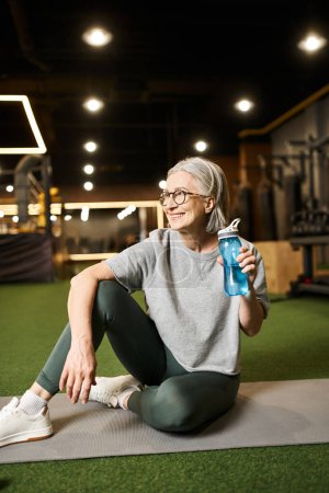 cheerful senior sportswoman in cozy t shirt sitting and holding water bottle and looking away