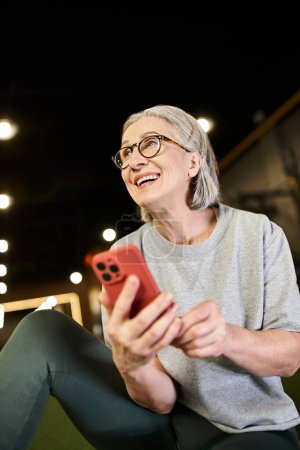 deportista madura alegre en camiseta gris con gafas sosteniendo teléfono inteligente y mirando hacia otro lado
