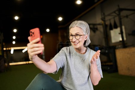 gut aussehende positive reife Sportlerin in kuscheliger Kleidung mit Brille beim Videoanruf im Fitnessstudio